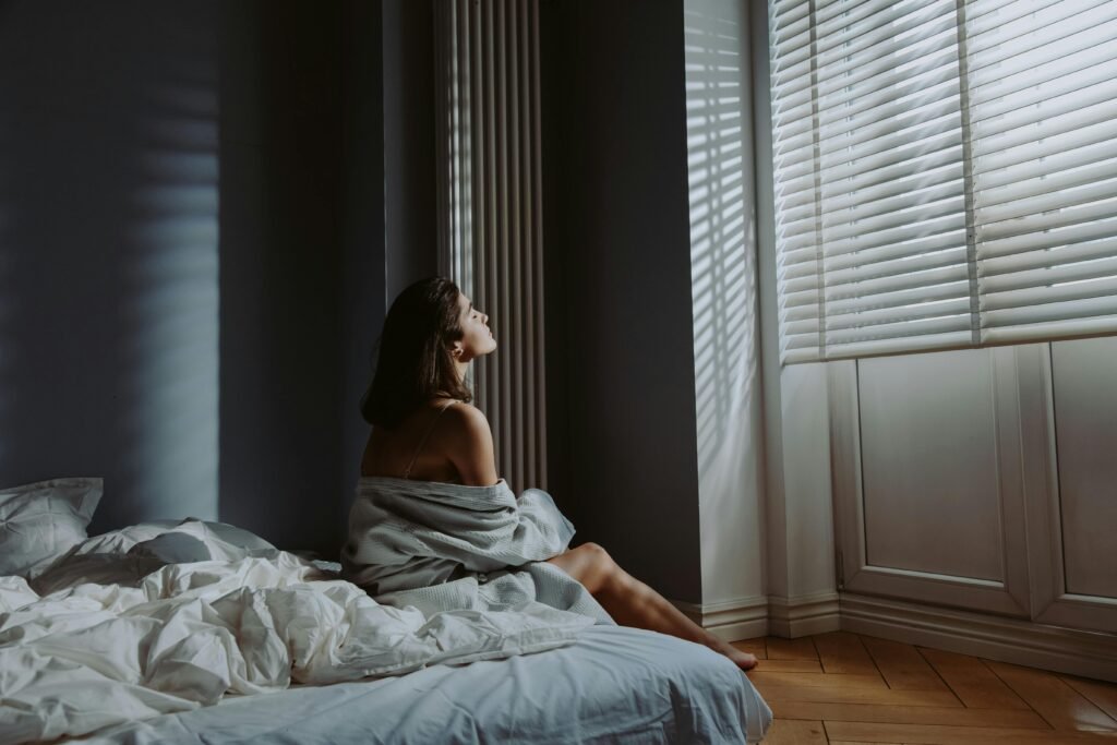 Woman sitting on bed in cozy bedroom, wrapped in a blanket with sunlight streaming through blinds.