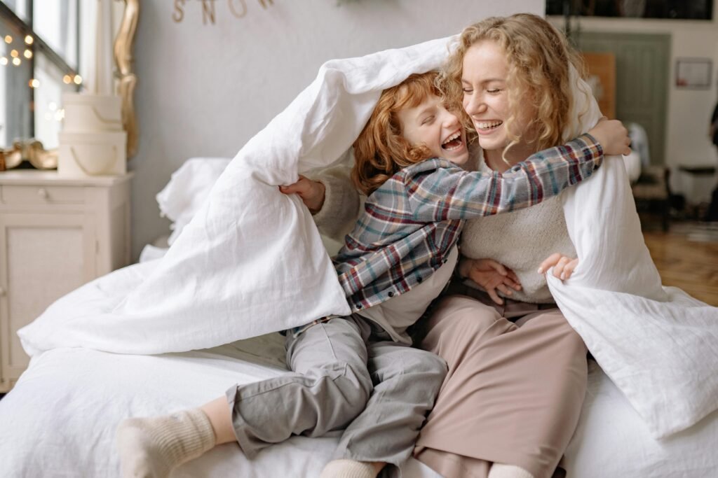 A mother and daughter share a joyful moment, wrapped in a cozy blanket at home.